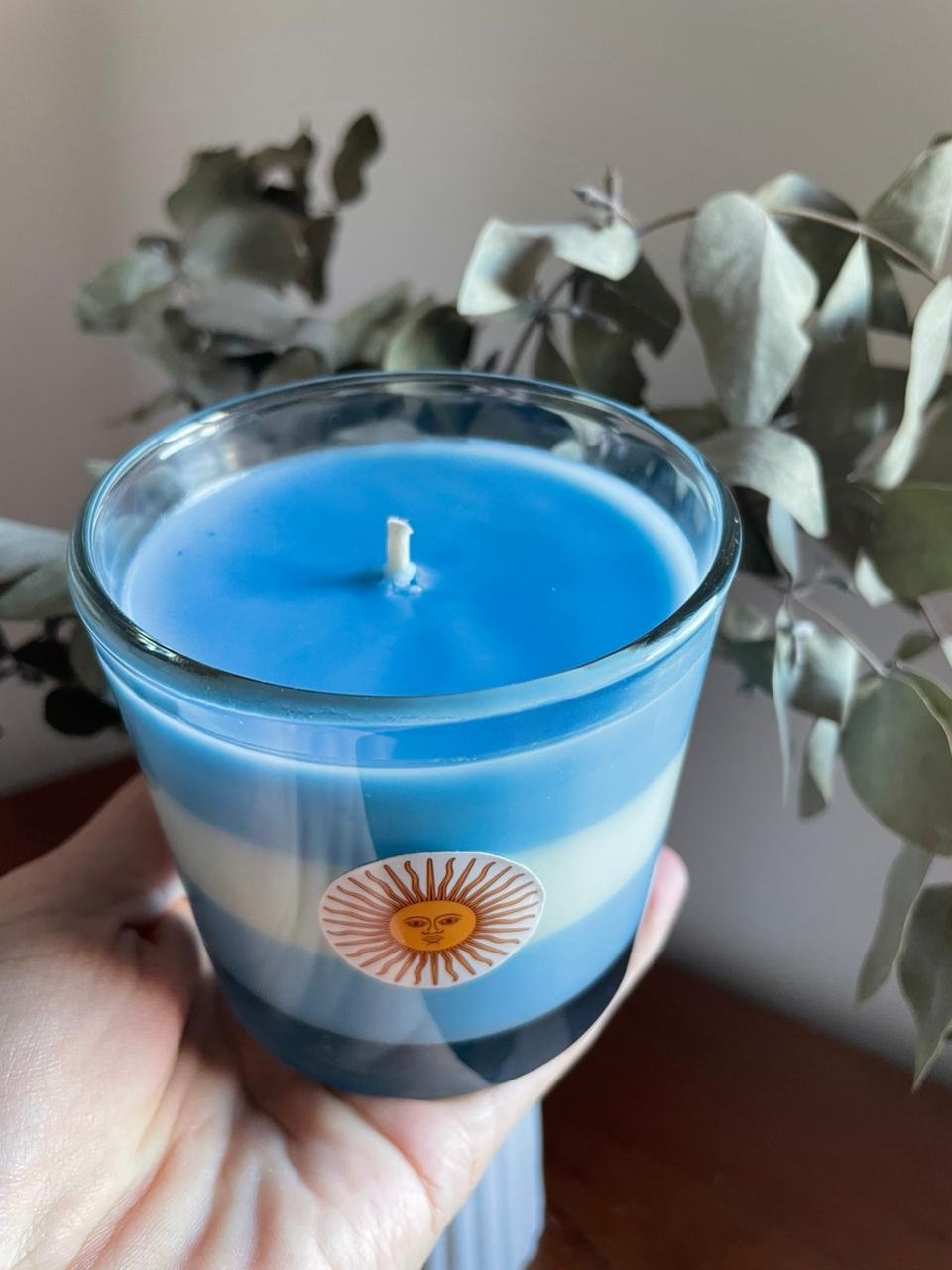 Vista superior de vela aromática celeste y blanca rodeada de hojas de eucalipto, ideal para regalo empresarial.